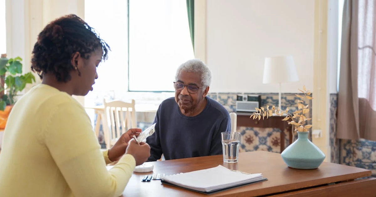 A hospice nurse with a clipboard speaking with an elderly patient and family member in a home living room