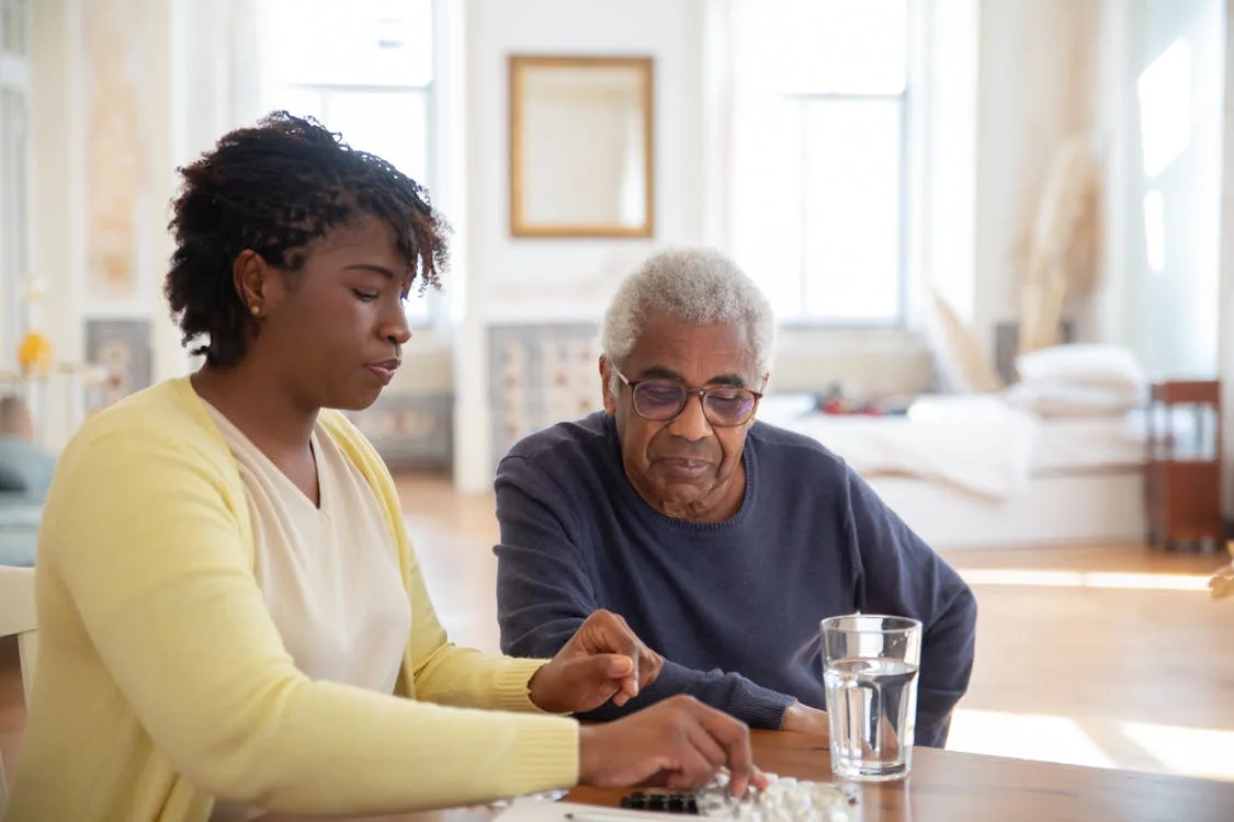 A caregiver sitting with an elderly man at a table, helping him sort his medications at home