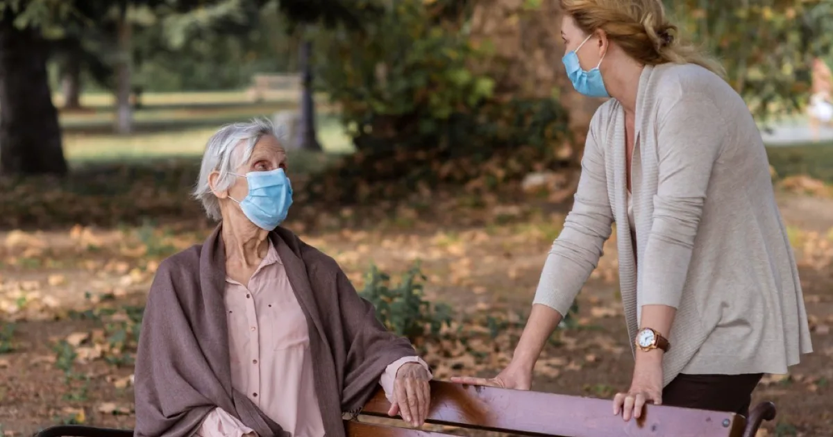 An elderly woman seated on a park bench wearing a face mask, talking with a younger caregiver standing beside her outdoors